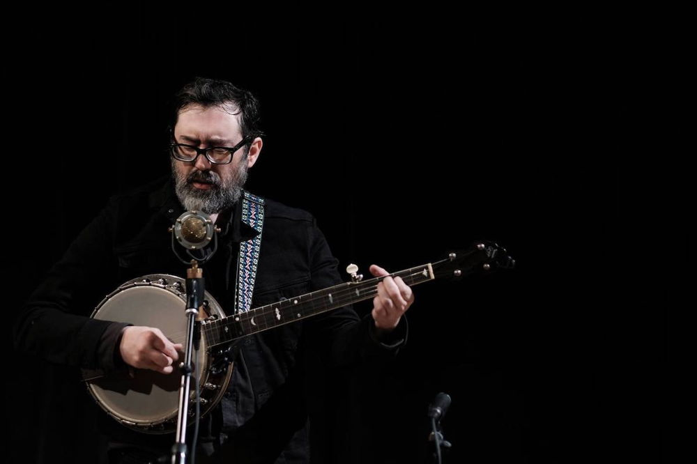 Brett wears a black shirt and stands against a black background, playing banjo and singing into a microphone. His hands are blurred from the motion of playing.