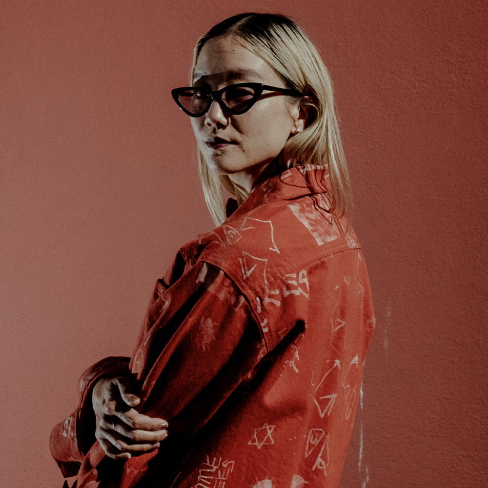 An Asian American woman poses with her body in profile, looking into the camera over her left shoulder. She is wearing a red denim jacket and black sunglasses with her blonde dyed hair around her shoulders, and she is standing against a red-orange painted house.