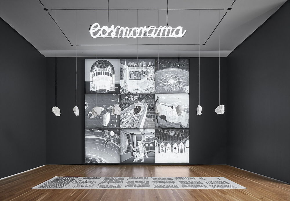 Photo of a gallery installation. A grid composed of nine retrofuturistic celestial images and diagrams is hung on the wall. White rocks hang from the ceiling in front of the grid, hovering over a long white paper with black text on it resting on the floor. A white neon sign reading “cosmorama” hangs from the ceiling.