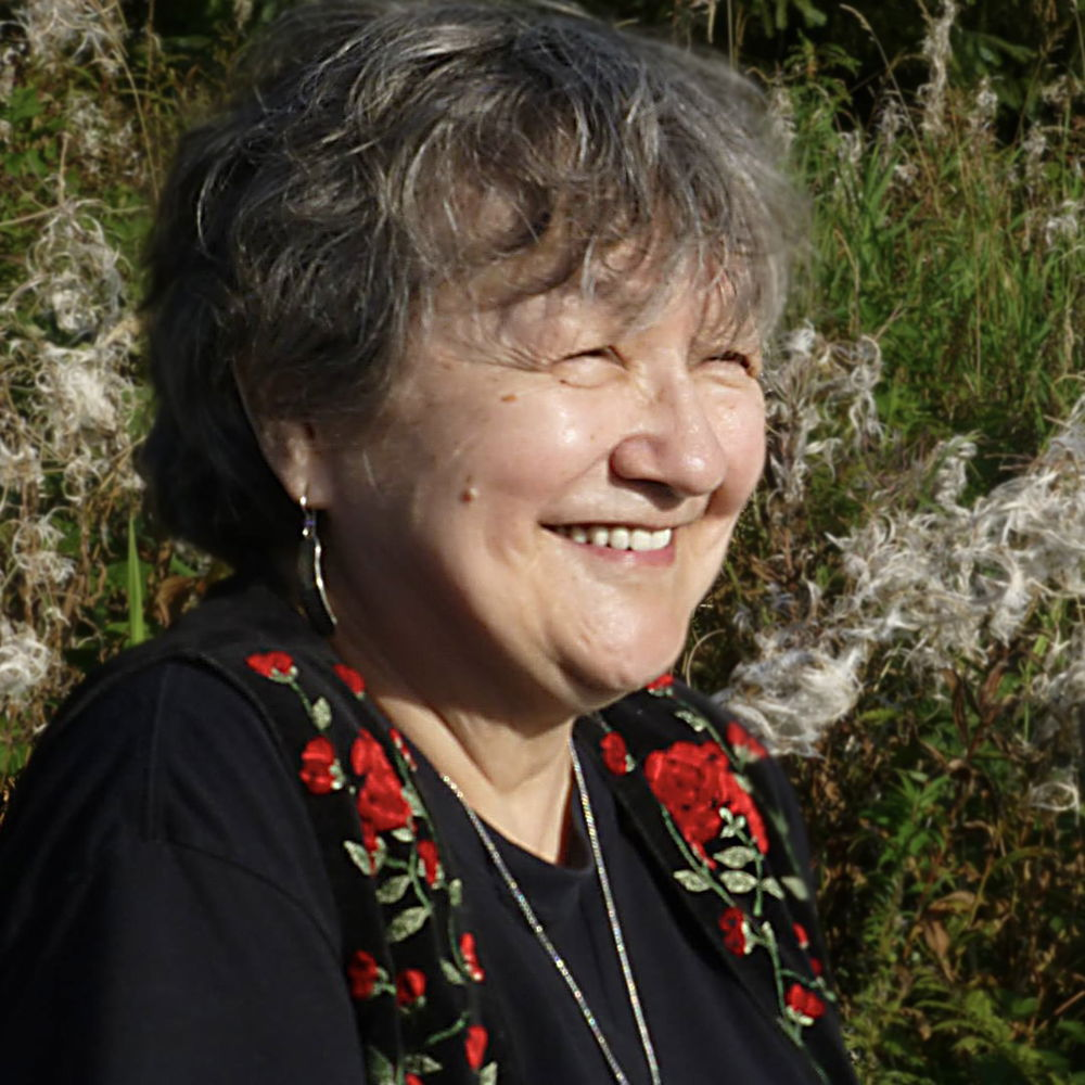 A person with short gray hair smiles warmly, their gaze off camera. They are wearing a flower-patterned vest, earrings, and a necklace and standing in front of a grassy backdrop.